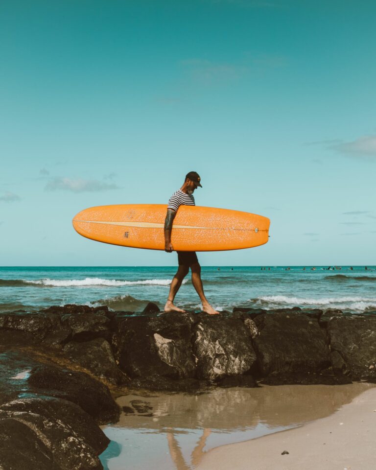 Surfer carrying an orange surfboard on a rocky beach