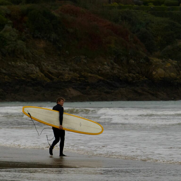 Surfer in a wetsuit carrying a yellow surfboard along the beach